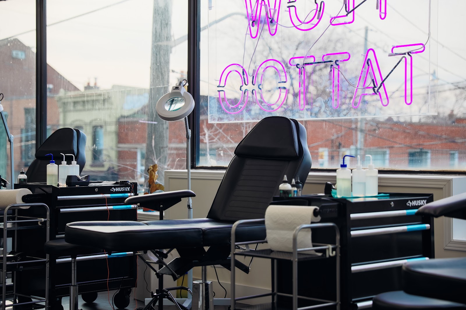 Inside the studio: black tattoo chairs and workstations by a big window with a pink neon tattoo sign